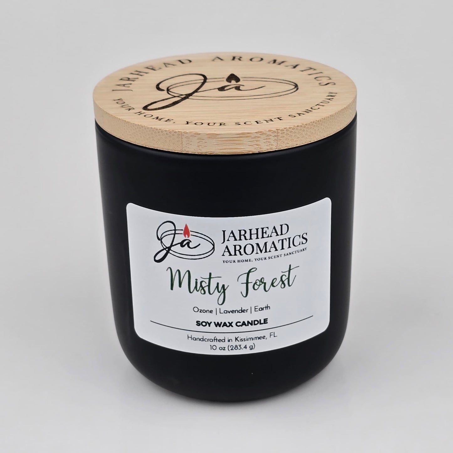 A 10 oz black soy wax candle in a jar with a wooden lid labeled “Jarhead Aromatics Misty Forest,” handcrafted in Kissimmee, FL. The Misty Forest Soy Wax Candle by Jarhead Aromatics features earthy moss, lavender, and juniper scents for a soothing, nature-inspired fragrance.