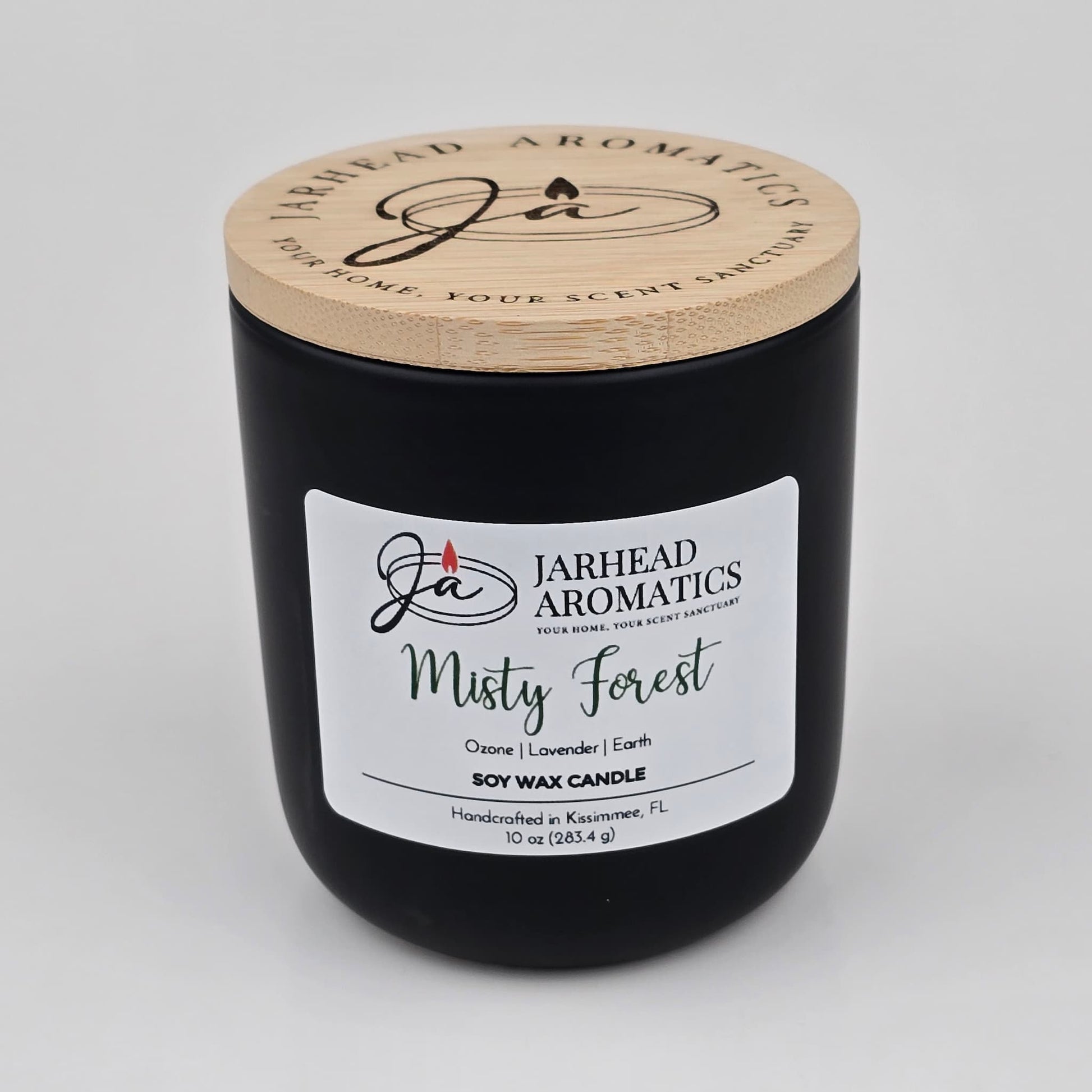 A 10 oz black soy wax candle in a jar with a wooden lid labeled “Jarhead Aromatics Misty Forest,” handcrafted in Kissimmee, FL. The Misty Forest Soy Wax Candle by Jarhead Aromatics features earthy moss, lavender, and juniper scents for a soothing, nature-inspired fragrance.