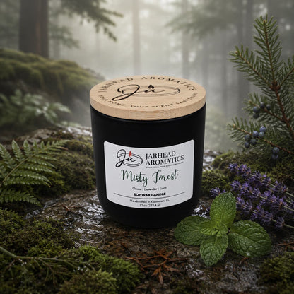 A black Jarhead Aromatics Misty Forest Soy Wax Candle rests on a bed of moss, surrounded by lavender, juniper, and pine branches, with a misty forest in the background. This candle combines natural soy wax with earthy moss, lavender, and juniper scents to bring the tranquil aroma of the woods into your space.