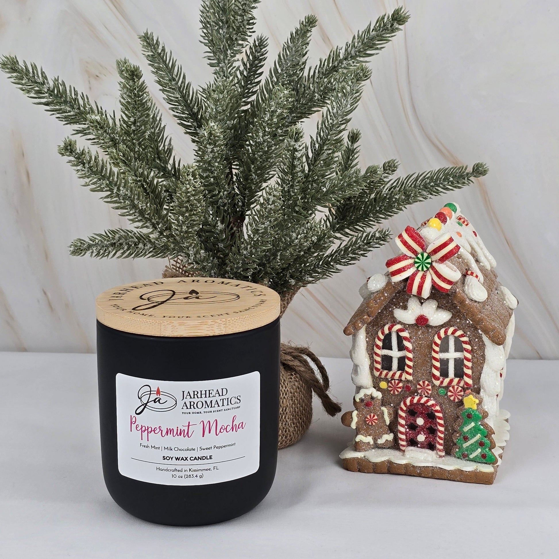 A black Peppermint Mocha - Soy Wax Candle from Jarhead Aromatics’ Winter Collection sits near a small artificial pine tree and gingerbread house on a light marble background, filling the air with a rich chocolate scent.