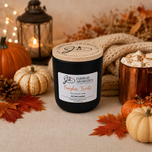 A black Jarhead Aromatics Pumpkin Treats Soy Wax Candle, featuring pumpkin caramel, maple pecan, and vanilla fragrances, sits on a beige cloth surrounded by pumpkins, autumn leaves, a knit blanket, fairy lights, and a mug of marshmallows—creating a cozy fall scene filled with the warm glow and inviting scent of this autumn candle.