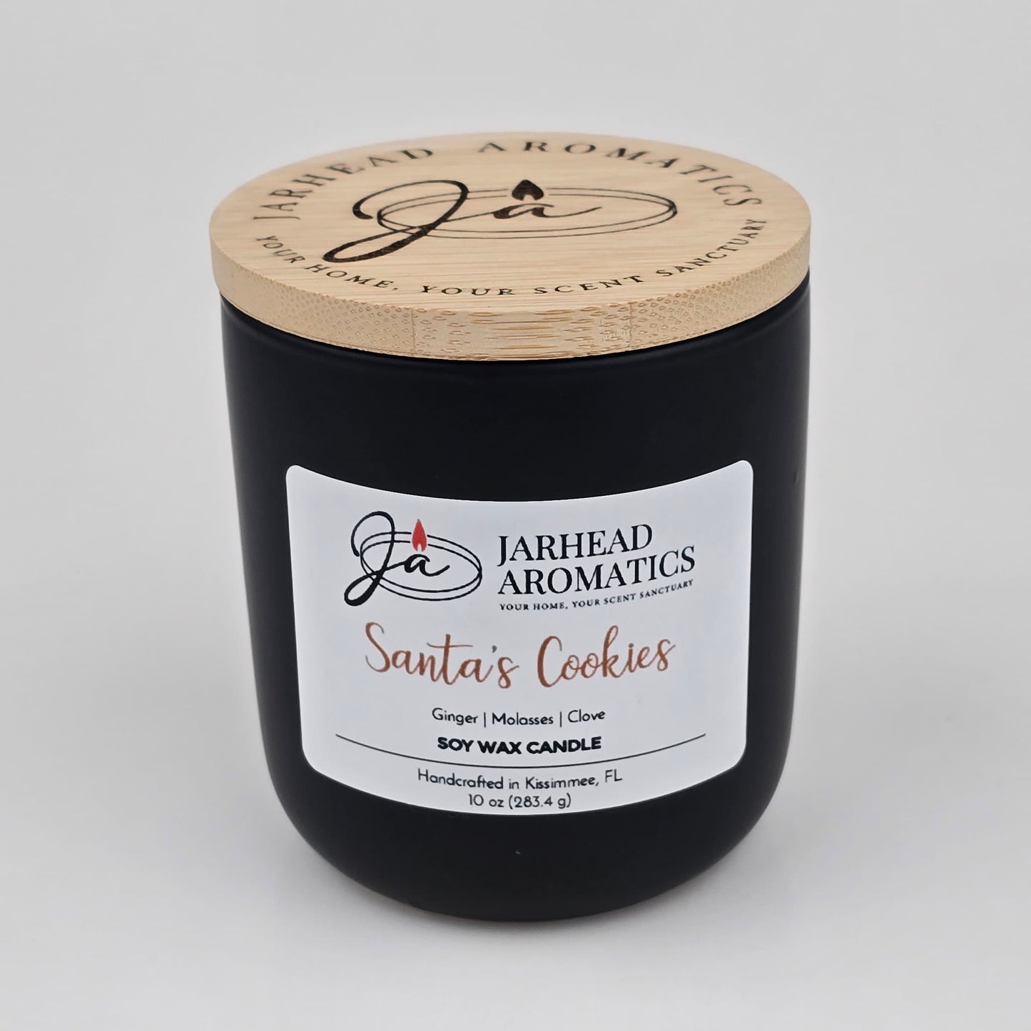 A black candle jar with a wooden lid from Jarhead Aromatics, labeled "Santa’s Cookies Soy Wax Candle | Ginger, Brown Sugar, Molasses & Vanilla Holiday Candle." Handcrafted in Kissimmee, FL, this soy wax candle features the festive scent of ginger, brown sugar, molasses, and vanilla—making it the perfect holiday candle to bring cozy warmth to any space.