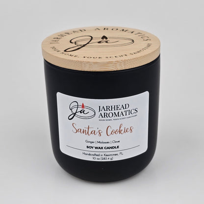 A black candle jar with a wooden lid from Jarhead Aromatics, labeled "Santa’s Cookies Soy Wax Candle | Ginger, Brown Sugar, Molasses & Vanilla Holiday Candle." Handcrafted in Kissimmee, FL, this soy wax candle features the festive scent of ginger, brown sugar, molasses, and vanilla—making it the perfect holiday candle to bring cozy warmth to any space.