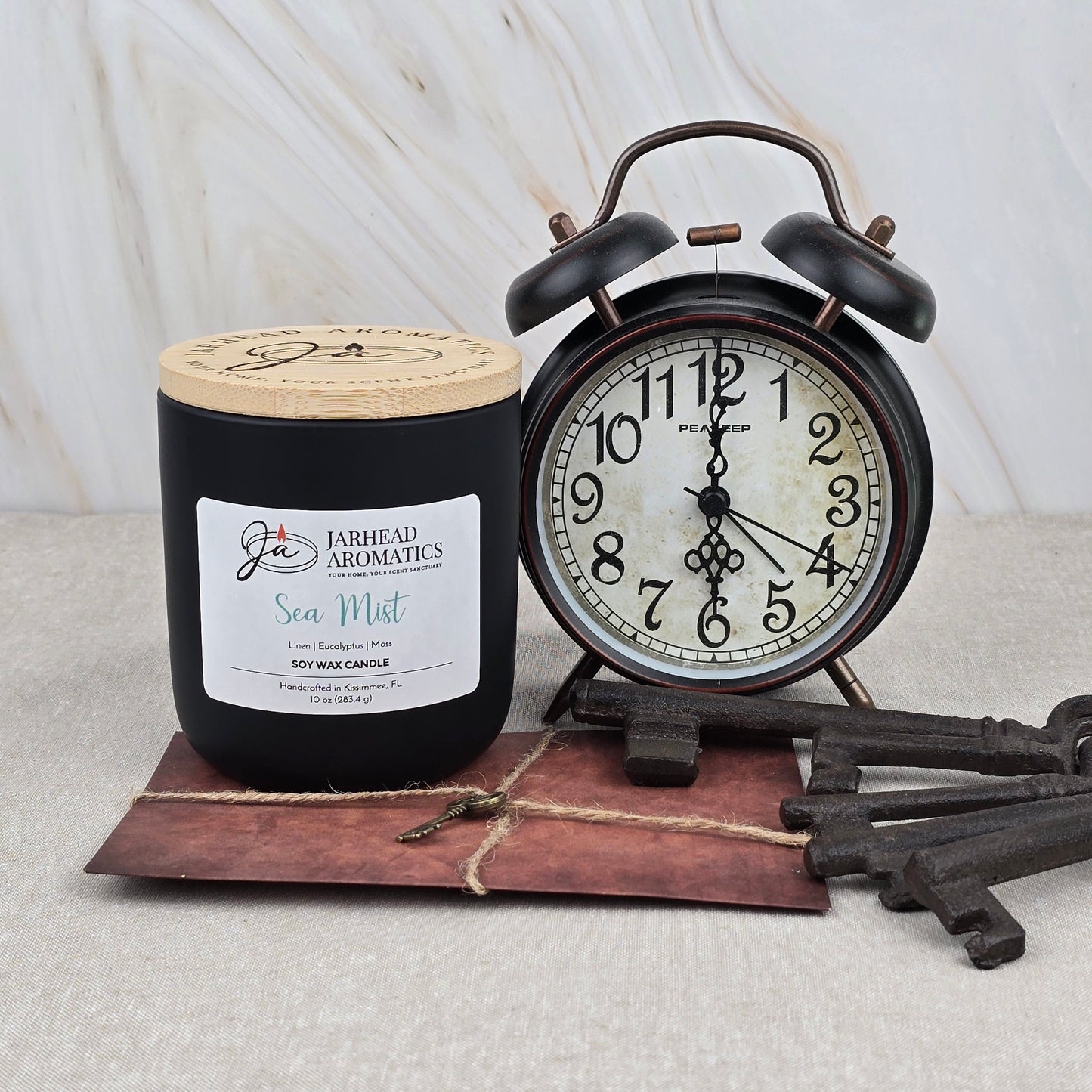 A Sea Mist Soy Wax Candle | Clean Coastal Linen & Sea Salt by Jarhead Aromatics sits beside a vintage black alarm clock and two decorative keys on a brown envelope with string, all arranged on a light marble background.