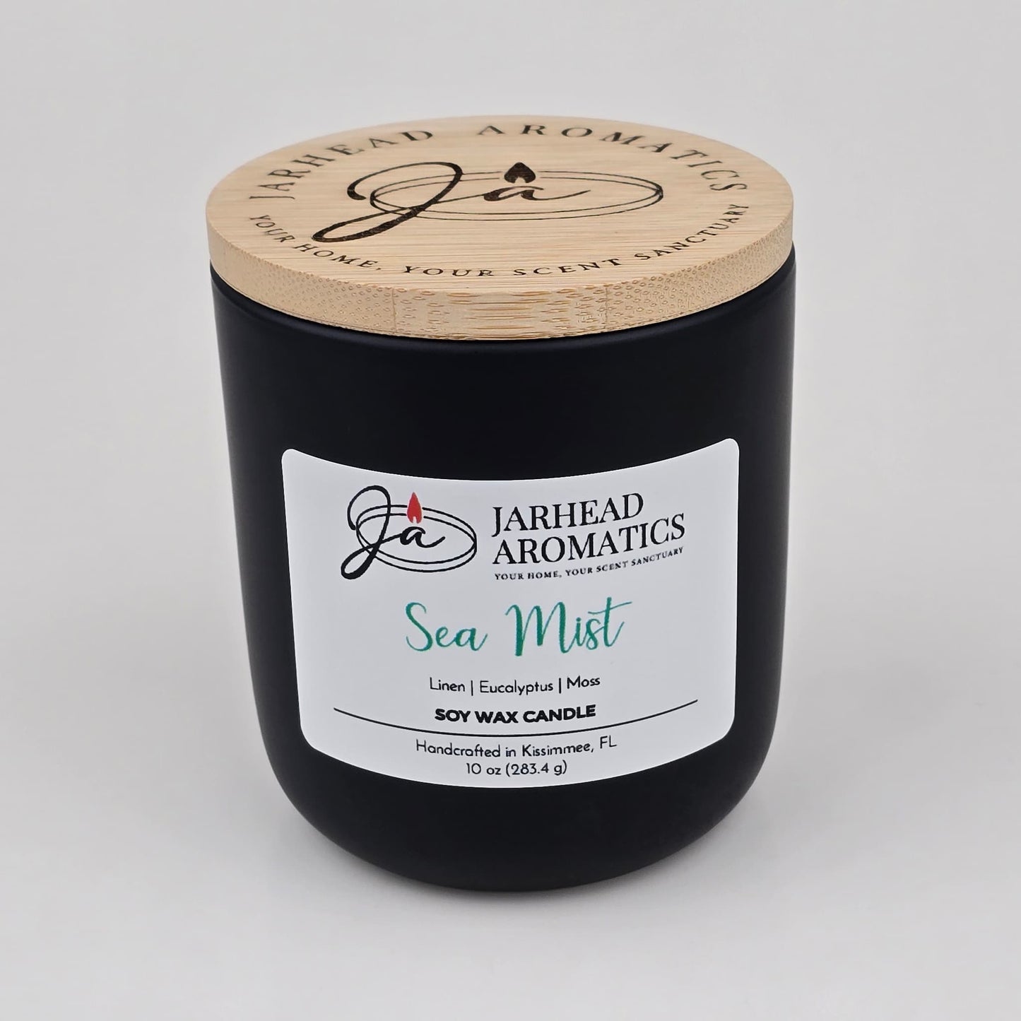 A black candle jar with a wooden lid from Jarhead Aromatics, labeled Sea Mist Soy Wax Candle | Clean Coastal Linen & Sea Salt Candle, featuring an invigorating aquatic floral fragrance of linen, eucalyptus, and moss. This coastal candle is handcrafted in Kissimmee, FL and contains 10 oz (283.4 g).