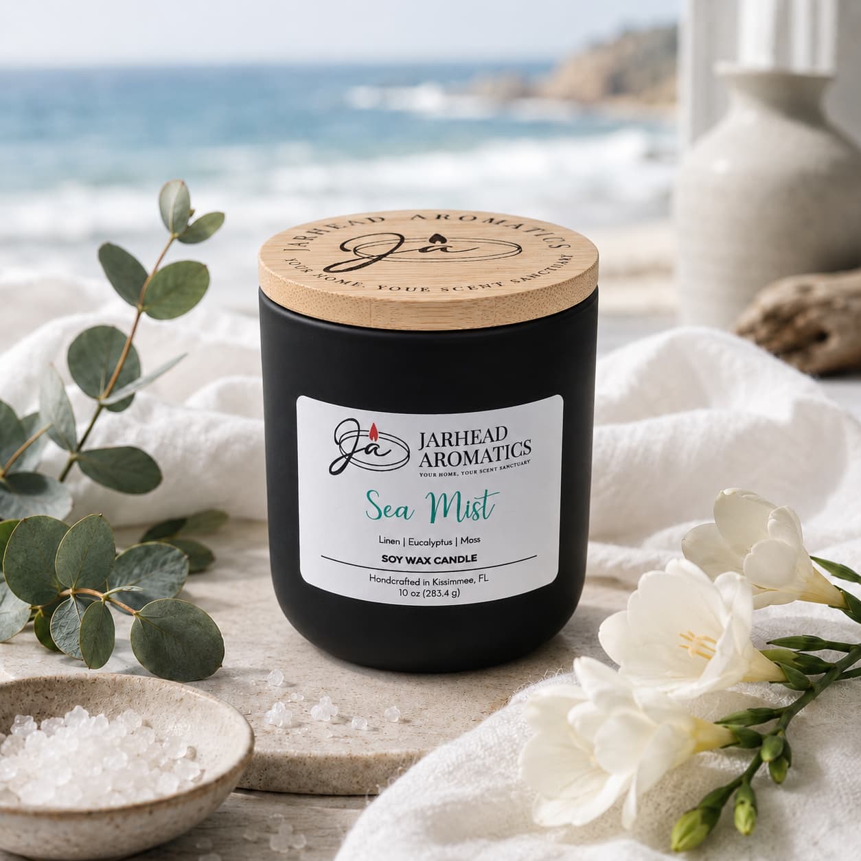 A Jarhead Aromatics Sea Mist Soy Wax Candle | Clean Coastal Linen & Sea Salt Candle sits on a table alongside sea salt, eucalyptus leaves, and white flowers. The blurred ocean and cliffs in the background evoke a tranquil coastal vibe. Infusing the air with an aquatic floral scent, this candle perfectly embodies the refreshing essence of seaside linen and salty breezes.