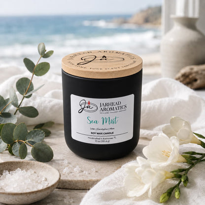A Jarhead Aromatics Sea Mist Soy Wax Candle | Clean Coastal Linen & Sea Salt Candle sits on a table alongside sea salt, eucalyptus leaves, and white flowers. The blurred ocean and cliffs in the background evoke a tranquil coastal vibe. Infusing the air with an aquatic floral scent, this candle perfectly embodies the refreshing essence of seaside linen and salty breezes.