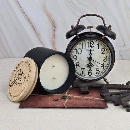 A black vintage alarm clock, the Misty Forest soy wax candle by Jarhead Aromatics, and old-fashioned keys sit on a brown leather pouch against a light marble background.