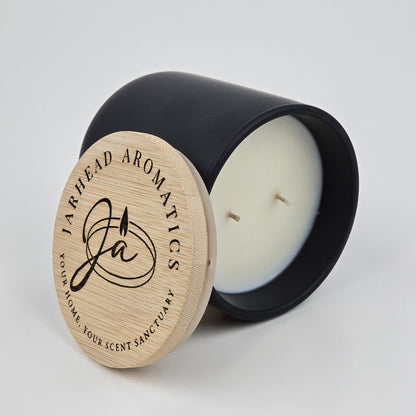 A black, two-wick candle—The Lumberjack Soy Wax Candle by Jarhead Aromatics—rests beside its engraved wooden lid, exuding cedar, oak, and mahogany notes for a masculine woodland cabin scent.
