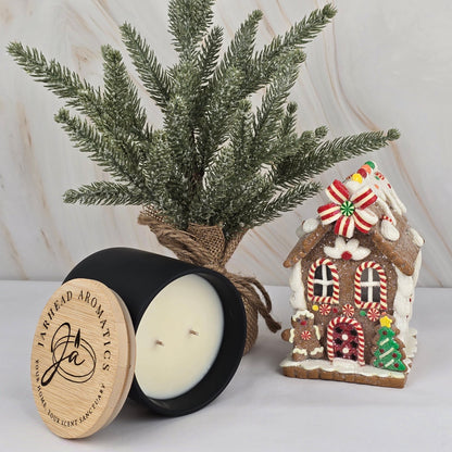 A Soy Wax Candle by Jarhead Aromatics, in a black jar with wooden lid, sits next to a faux pine and gingerbread house on a white surface with a marble background.