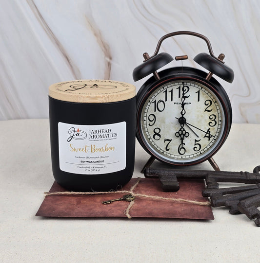 A black Jarhead Aromatics Sweet Bourbon - Soy Wax Candle, celebrated for its rich scent, rests alongside a vintage black alarm clock on a twine-tied brown leather journal with old keys, set against a marble-patterned backdrop.