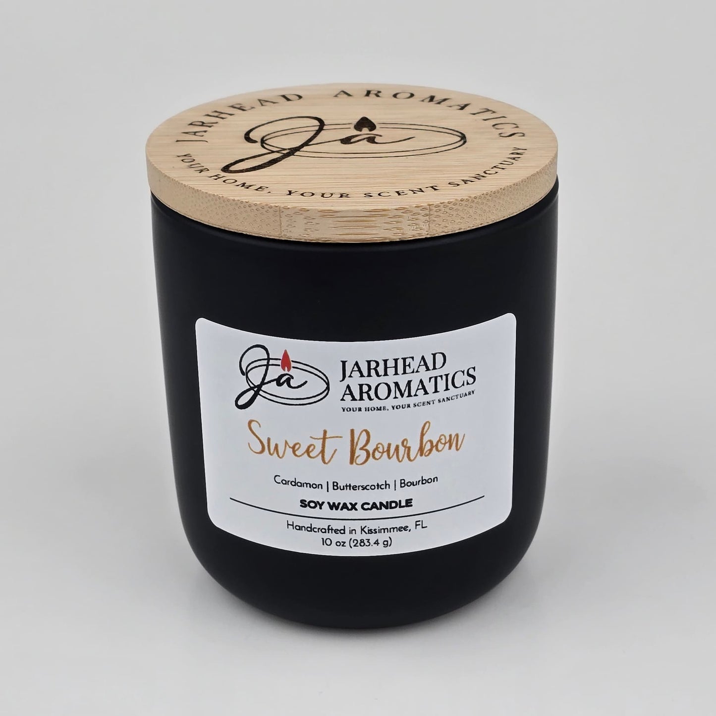 A black candle jar with a wooden lid features the Jarhead Aromatics label for their Sweet Bourbon Soy Wax Candle. The label highlights warm bourbon, brown sugar, and oak notes. This hand-poured soy candle is handcrafted in Kissimmee, FL, and offers a long-lasting fragrance perfect for those who appreciate the inviting aroma of a Warm Bourbon, Brown Sugar & Oak Candle.