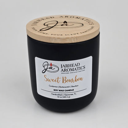 A black candle jar with a wooden lid features the Jarhead Aromatics label for their Sweet Bourbon Soy Wax Candle. The label highlights warm bourbon, brown sugar, and oak notes. This hand-poured soy candle is handcrafted in Kissimmee, FL, and offers a long-lasting fragrance perfect for those who appreciate the inviting aroma of a Warm Bourbon, Brown Sugar & Oak Candle.