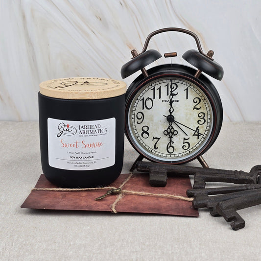 A Jarhead Aromatics Sweet Sunrise Soy Wax Candle, featuring bright citrus, agave & peach notes, sits beside a vintage alarm clock showing 11:02. Three antique keys and a brown envelope are on the white marbled surface.