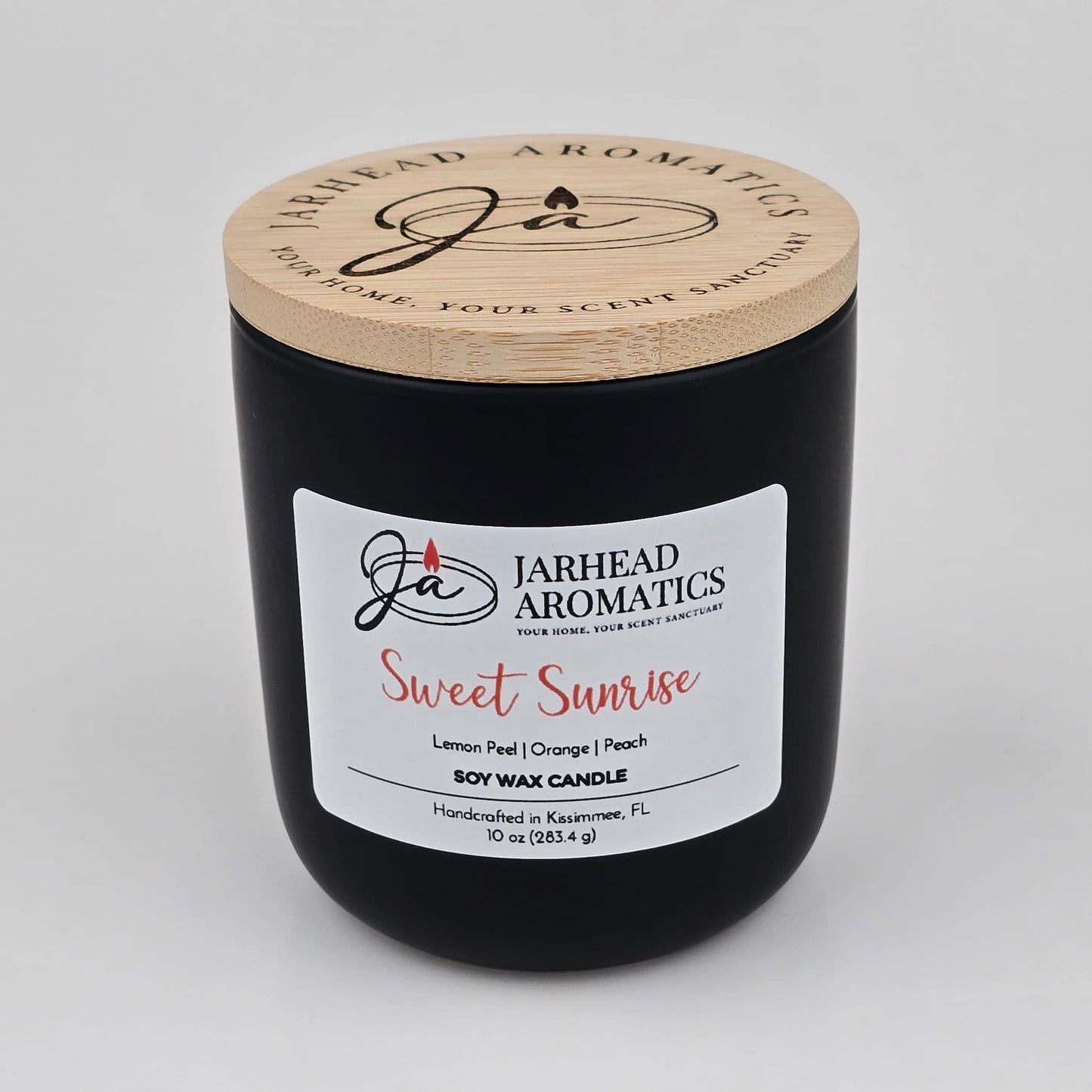 A 10 oz black candle jar with a wooden lid, labeled Jarhead Aromatics Sweet Sunrise Soy Wax Candle, handcrafted in Kissimmee, FL. This candle features bright citrus, agave, and peach fragrance notes for an invigorating scent experience.