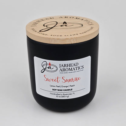 A 10 oz black candle jar with a wooden lid, labeled Jarhead Aromatics Sweet Sunrise Soy Wax Candle, handcrafted in Kissimmee, FL. This candle features bright citrus, agave, and peach fragrance notes for an invigorating scent experience.