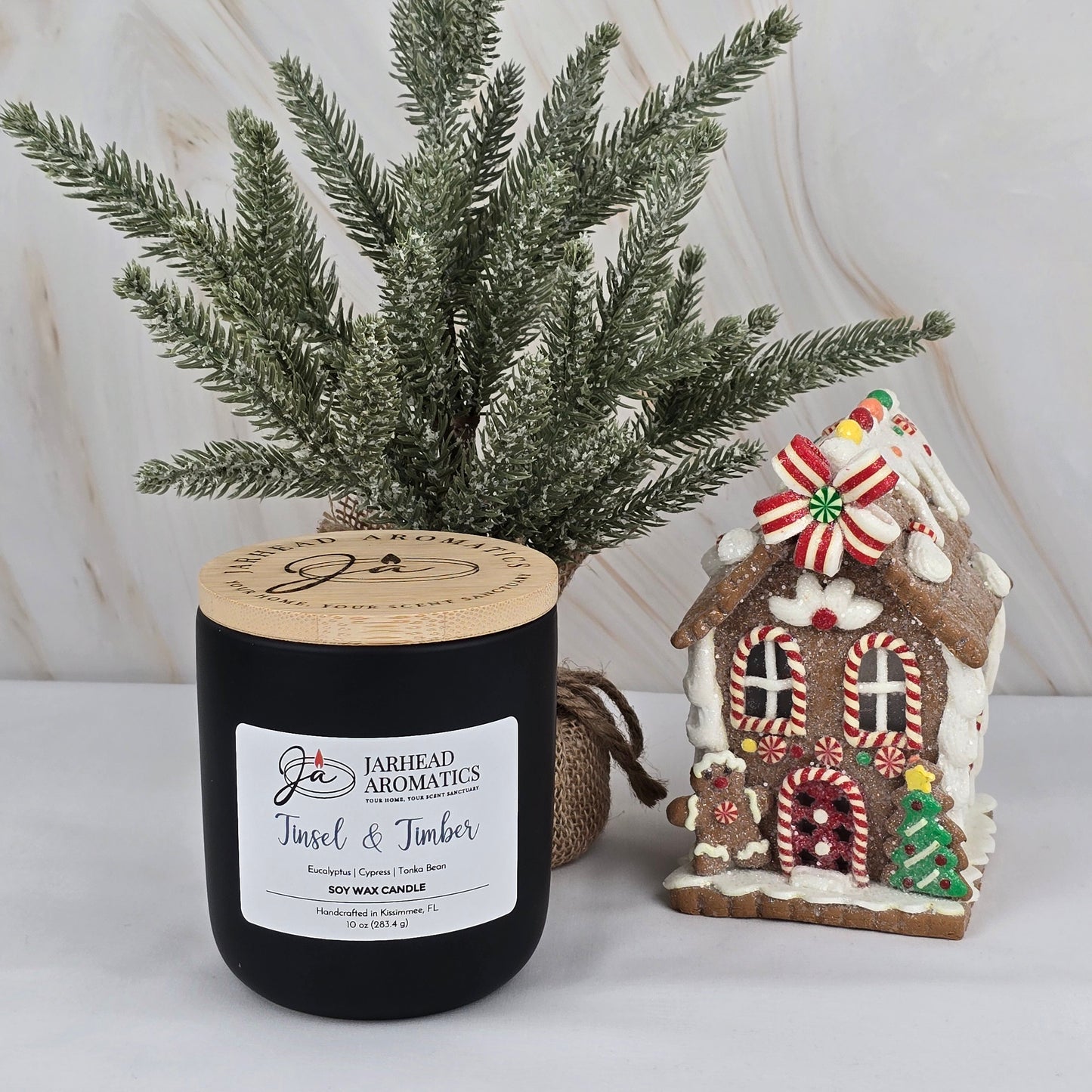 A Jarhead Aromatics Tinsel & Timber Soy Wax Candle from the Winter Collection sits on a white surface beside a small artificial pine tree and a decorative gingerbread house with colorful candy accents.
