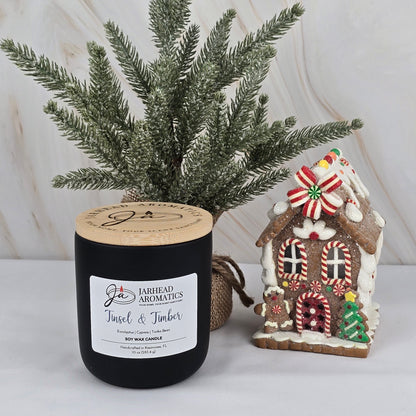 A Jarhead Aromatics Tinsel & Timber Soy Wax Candle from the Winter Collection sits on a white surface beside a small artificial pine tree and a decorative gingerbread house with colorful candy accents.