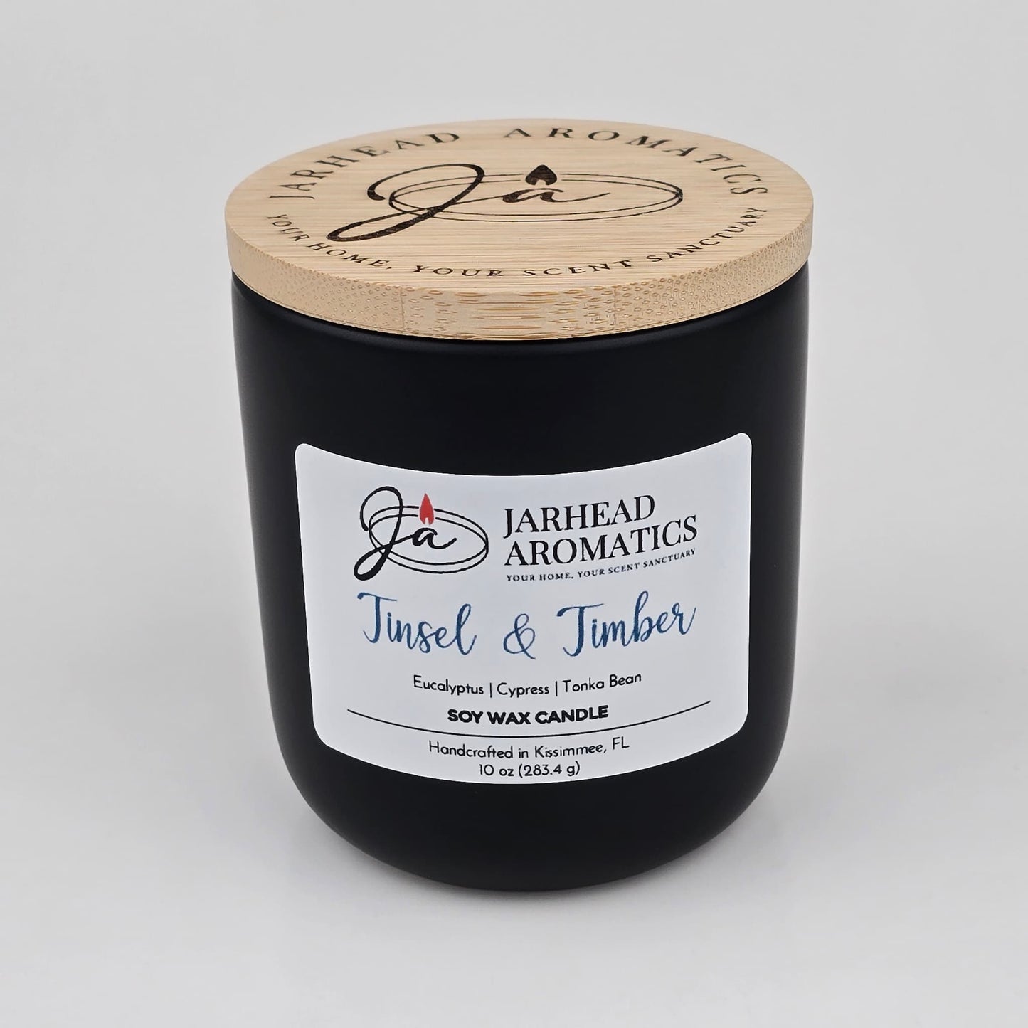 A black soy wax candle jar with a wooden lid from Jarhead Aromatics features a white label reading “Tinsel & Timber” and highlights notes of eucalyptus, pine, smoke, and tonka. Handmade in Kissimmee, FL, this inviting holiday candle brings an evergreen fragrance to any home.