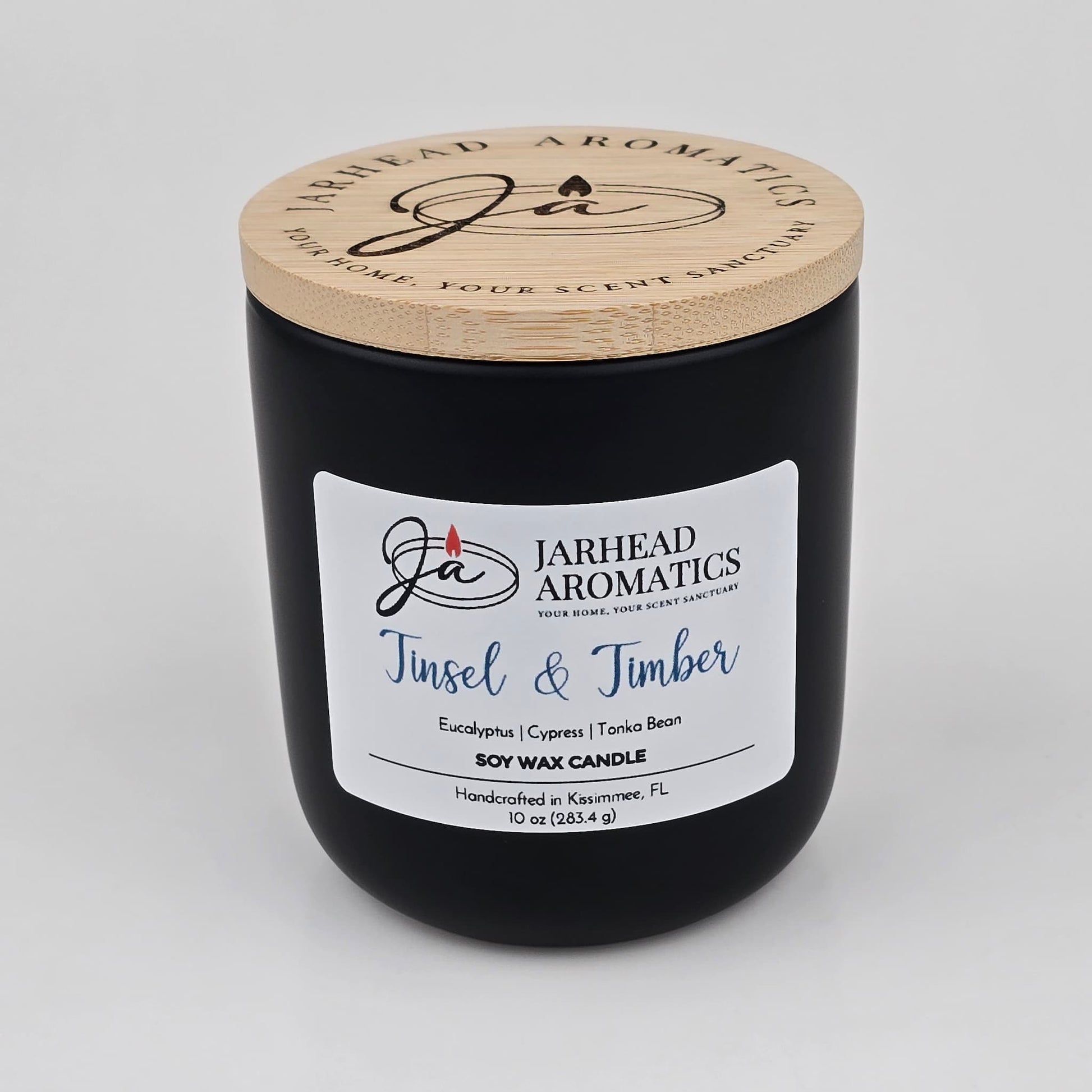 A black soy wax candle jar with a wooden lid from Jarhead Aromatics features a white label reading “Tinsel & Timber” and highlights notes of eucalyptus, pine, smoke, and tonka. Handmade in Kissimmee, FL, this inviting holiday candle brings an evergreen fragrance to any home.