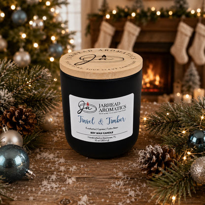 A Jarhead Aromatics Tinsel & Timber Soy Wax Candle, featuring eucalyptus, pine, smoke, and tonka notes, rests on a wooden table surrounded by pine branches and ornaments. A fireplace with stockings in the background completes the scene, infusing the room with a cozy holiday fragrance for a festive atmosphere.