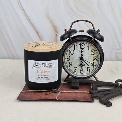 A black Jarhead Aromatics Tulip Bliss - Soy Wax Candle with essential oils sits by a vintage black alarm clock set to 9:10, three old metal keys, and a brown envelope, all atop a light marble surface.
