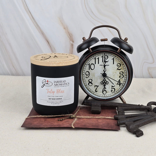 A black Jarhead Aromatics Tulip Bliss - Soy Wax Candle with essential oils sits by a vintage black alarm clock set to 9:10, three old metal keys, and a brown envelope, all atop a light marble surface.
