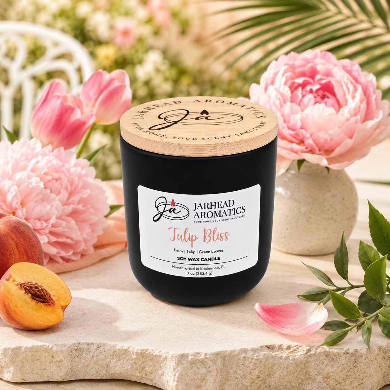 A black jar candle from Jarhead Aromatics, labeled "Tulip Bliss Soy Wax Candle | Peach, Tulip & Soft Musk Candle," rests on a stone surface surrounded by pink peonies, fresh peach slices, and green leaves. Sunlight streams through palm fronds in the background, illuminating this floral-scented candle and creating an inviting ambiance.
