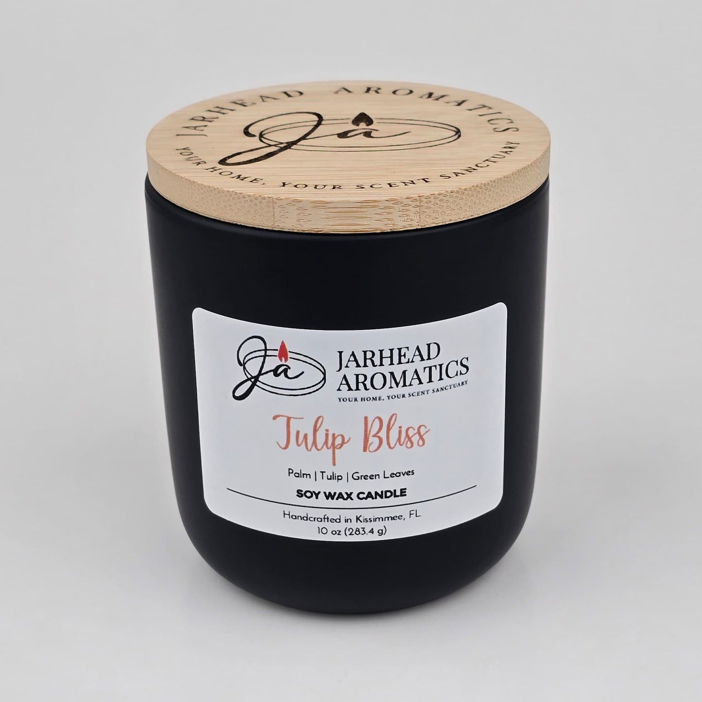 A black candle jar with a wooden lid labeled Jarhead Aromatics Tulip Bliss Soy Wax Candle rests on a white background. The label highlights notes of peach, tulip, and soft musk, describing it as a floral-scented soy wax candle handcrafted in Kissimmee, FL by Jarhead Aromatics.