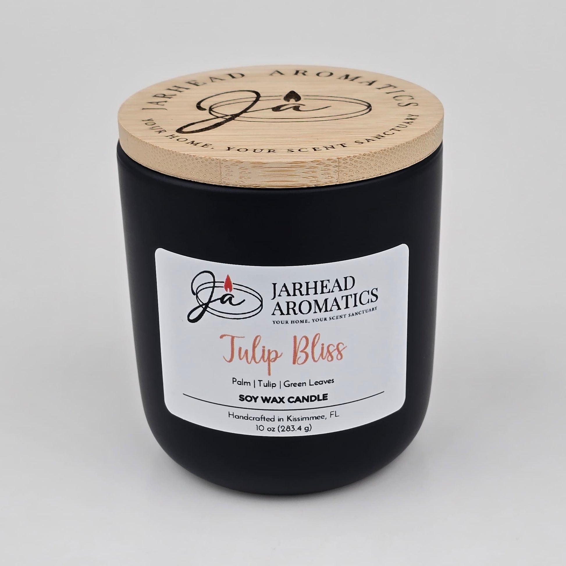 A black candle jar with a wooden lid labeled Jarhead Aromatics Tulip Bliss Soy Wax Candle rests on a white background. The label highlights notes of peach, tulip, and soft musk, describing it as a floral-scented soy wax candle handcrafted in Kissimmee, FL by Jarhead Aromatics.
