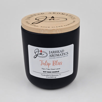A black candle jar with a wooden lid labeled Jarhead Aromatics Tulip Bliss Soy Wax Candle rests on a white background. The label highlights notes of peach, tulip, and soft musk, describing it as a floral-scented soy wax candle handcrafted in Kissimmee, FL by Jarhead Aromatics.