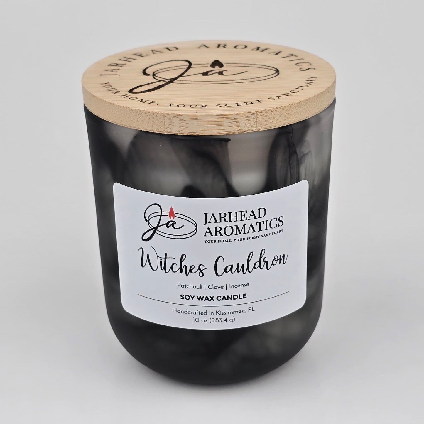 A handcrafted black glass candle jar with a wooden lid, labeled Jarhead Aromatics Witches Cauldron Soy Wax Candle, features notes of orange blossom, patchouli, clove, and incense; this Halloween candle is made in Kissimmee, FL.