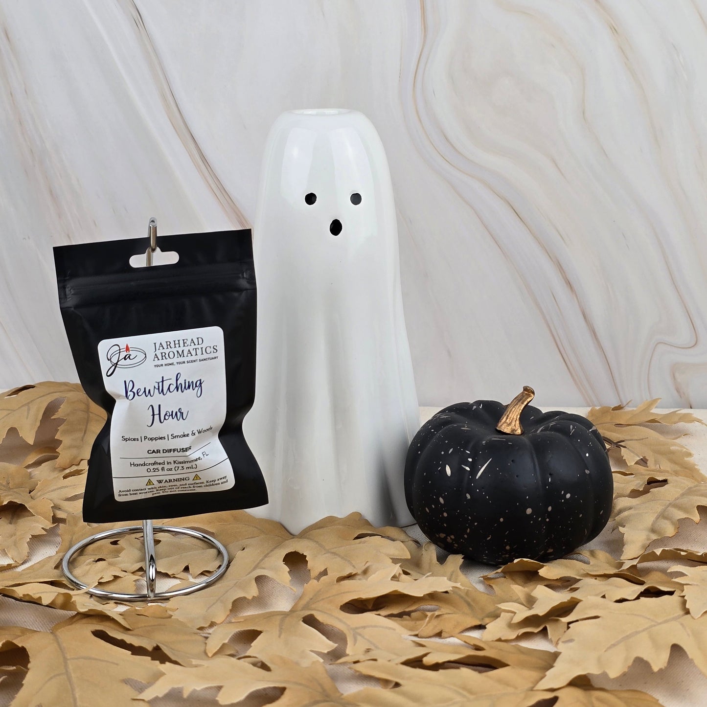 A white ceramic ghost, a black pumpkin, and Jarhead Aromatics Bewitching Hour - Car Diffuser from the Halloween Collection evoke a mystical aroma as they rest on dried leaves before a marble-patterned backdrop.