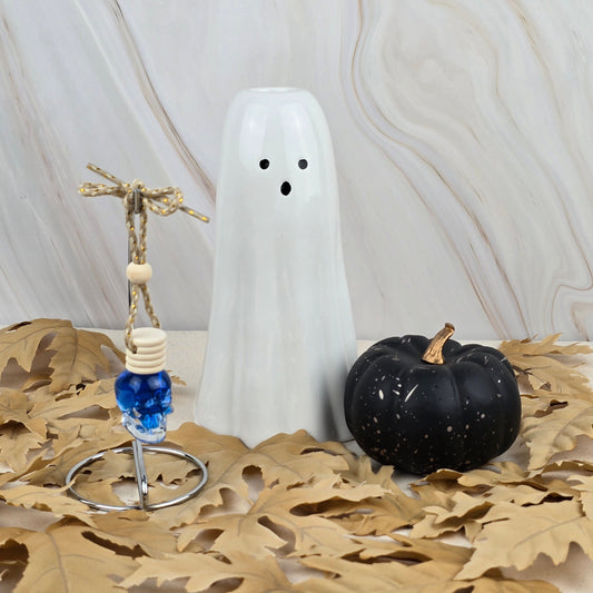 A white ceramic ghost, black pumpkin, and blue glass skull from the Bewitching Hour Car Diffuser by Jarhead Aromatics rest on tan leaves, capturing a mystical Halloween vibe against a marbled backdrop.