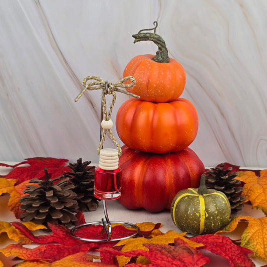A stack of three small orange pumpkins and a green pumpkin rest among pinecones and autumn leaves. In the foreground, Jarhead Aromatics Cran-Apple Delight Car Diffuser from the Fall Collection hangs, filled with red fragrance oil. Marble backdrop.