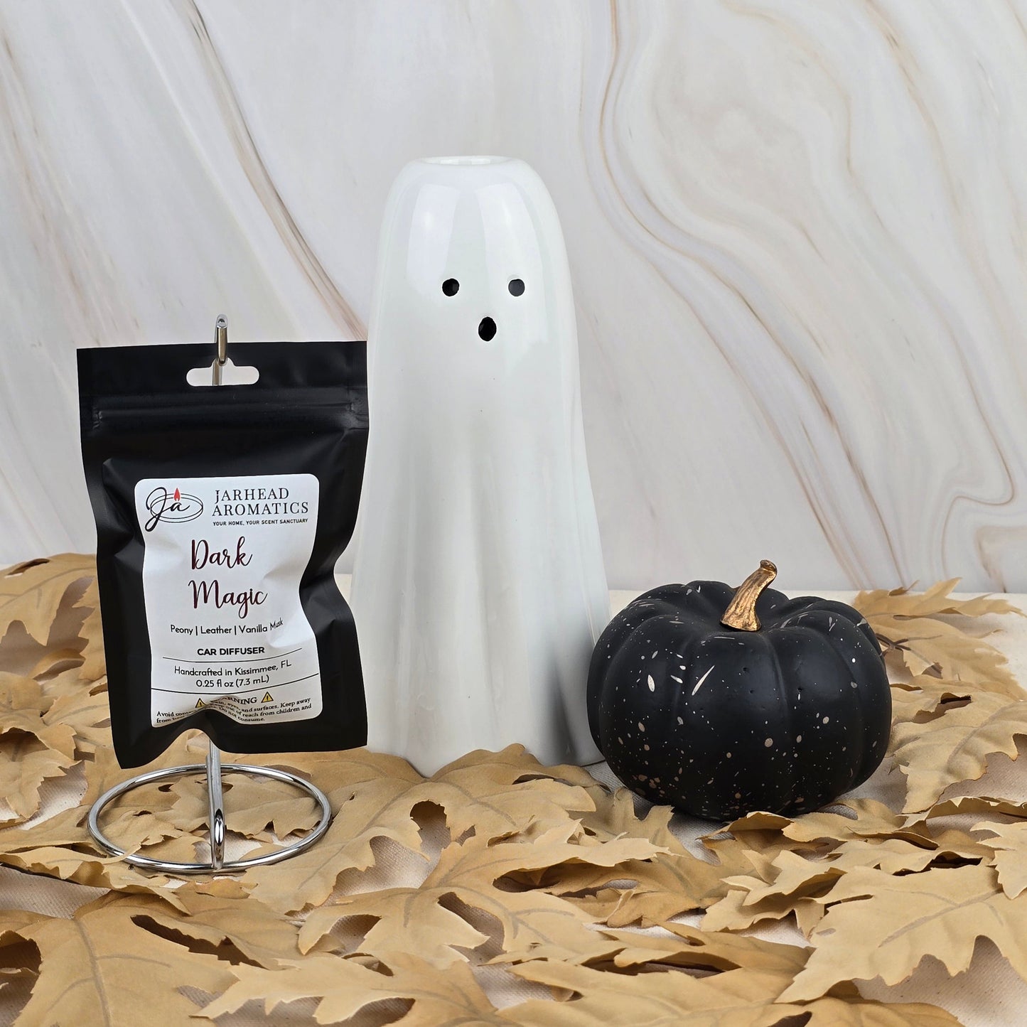 A ceramic ghost, a small black pumpkin, and Jarhead Aromatics Dark Magic Car Diffuser from the Halloween Collection rest on dry oak leaves against marble, creating a scene of mysterious allure and dark enchantment.