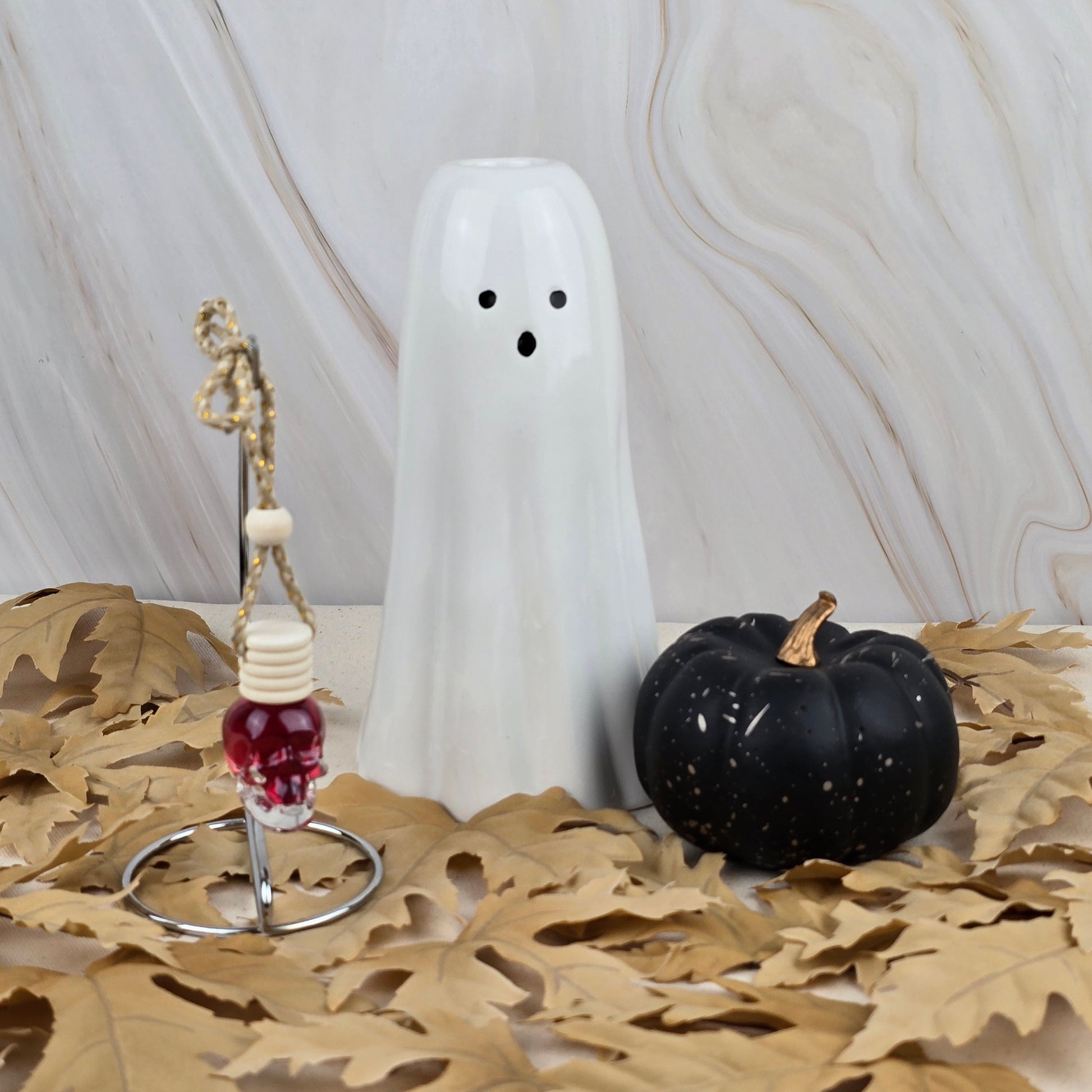 The Dark Magic Car Diffuser from Jarhead Aromatics’ Halloween Collection sits with a white ceramic ghost and black pumpkin on dried leaves, set against a marble-patterned background.
