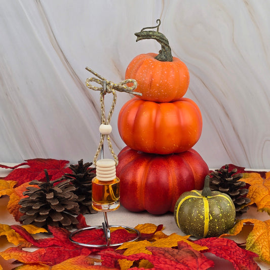A small stack of three orange pumpkins and one green pumpkin sits among autumn leaves and pinecones. In front, the Jarhead Aromatics Harvest Cream Car Diffuser from the Fall Collection hangs from a stand, adding festive fall fragrance.