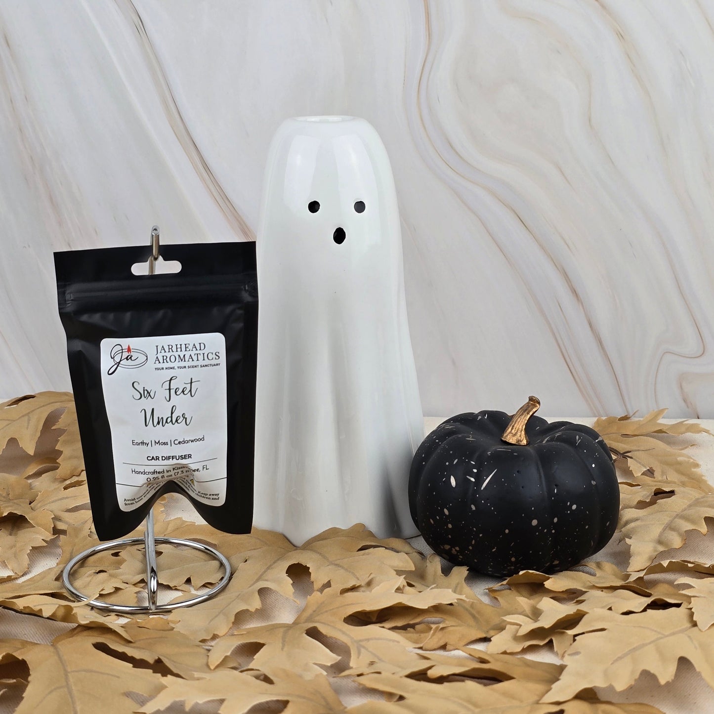 A white ceramic ghost, a black pumpkin with a gold stem, and Jarhead Aromatics Six Feet Under - Car Diffuser from the Halloween Collection are displayed on tan leaves against a marbled beige background.