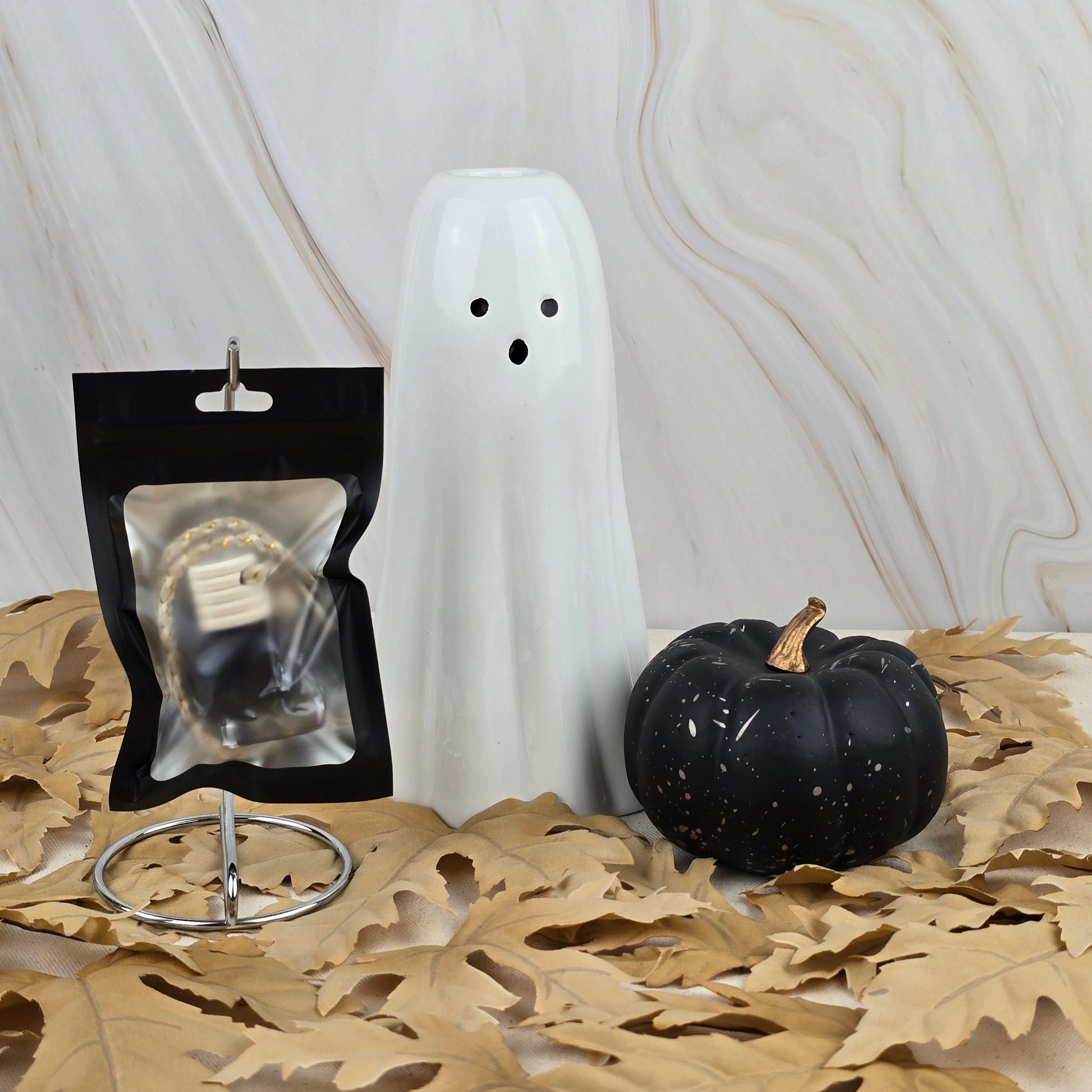A Witches Cauldron car diffuser from Jarhead Aromatics’ Halloween Collection sits on dried leaves beside ghost and pumpkin decor, with a string light, all set against marble.