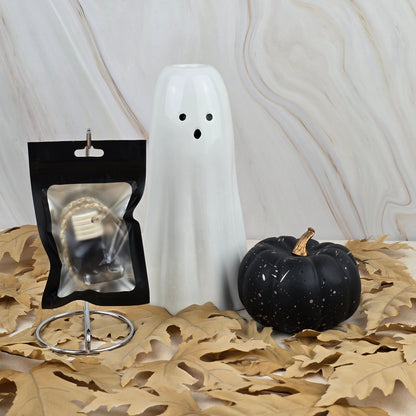 A Witches Cauldron car diffuser from Jarhead Aromatics’ Halloween Collection sits on dried leaves beside ghost and pumpkin decor, with a string light, all set against marble.