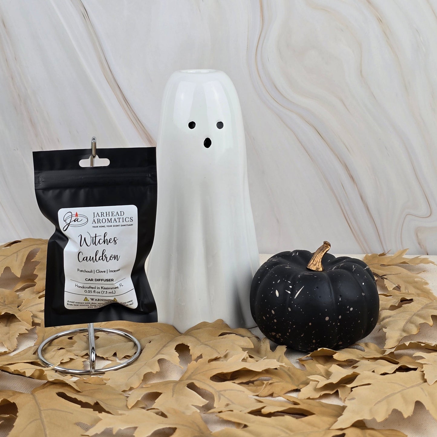 A white ceramic ghost, a black pumpkin, and Jarhead Aromatics Witches Cauldron Car Diffuser from the Halloween Collection are displayed on dried leaves against a marbled background.