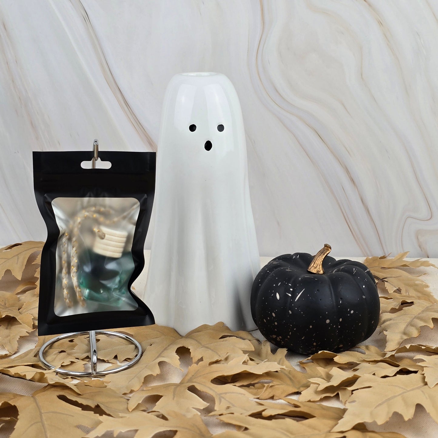 A white ceramic ghost, a black pumpkin with a gold stem, and a jewelry pouch on a stand sit among dried leaves against marble, evoking the haunting aroma of Jarhead Aromatics’ Zombie Queen Car Diffuser from the Halloween Collection.