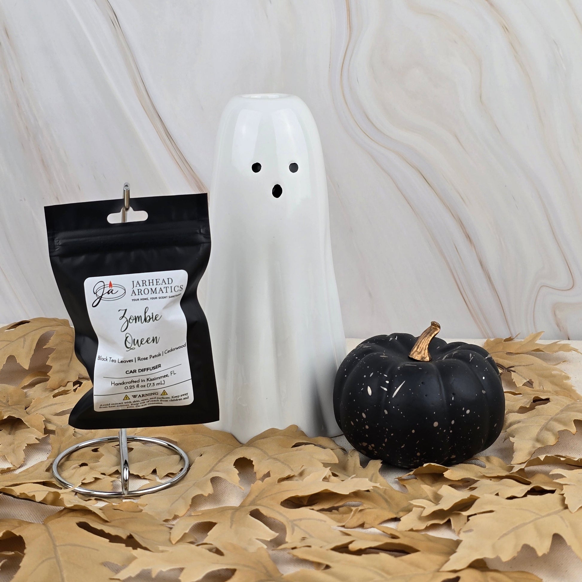 A ceramic ghost, a black pumpkin, and Jarhead Aromatics’ Zombie Queen Car Diffuser from the Halloween Collection rest on dried leaves over marble, evoking a spooky seasonal scent perfect for your car.