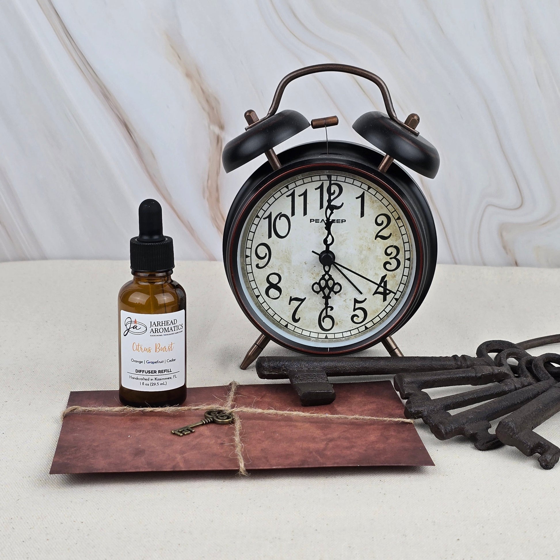A Citrus Burst Car Diffuser Refill by Jarhead Aromatics rests on a light surface with a marble-patterned background, accompanied by a vintage alarm clock, brown string-tied envelope, and old metal keys.
