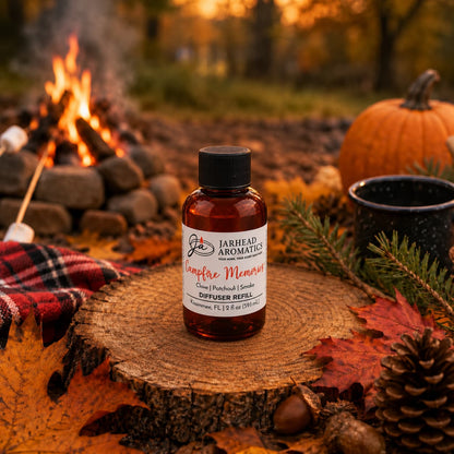 A bottle of Jarhead Aromatics Campfire Memories Diffuser Refill | Maple, Clove & Smoky Embers rests on a tree stump amid autumn leaves, pinecones, a pumpkin, and a mug, while a blurred campfire with roasting marshmallows flickers in the background—evoking the essence of home fragrance with an inviting autumn aroma.
