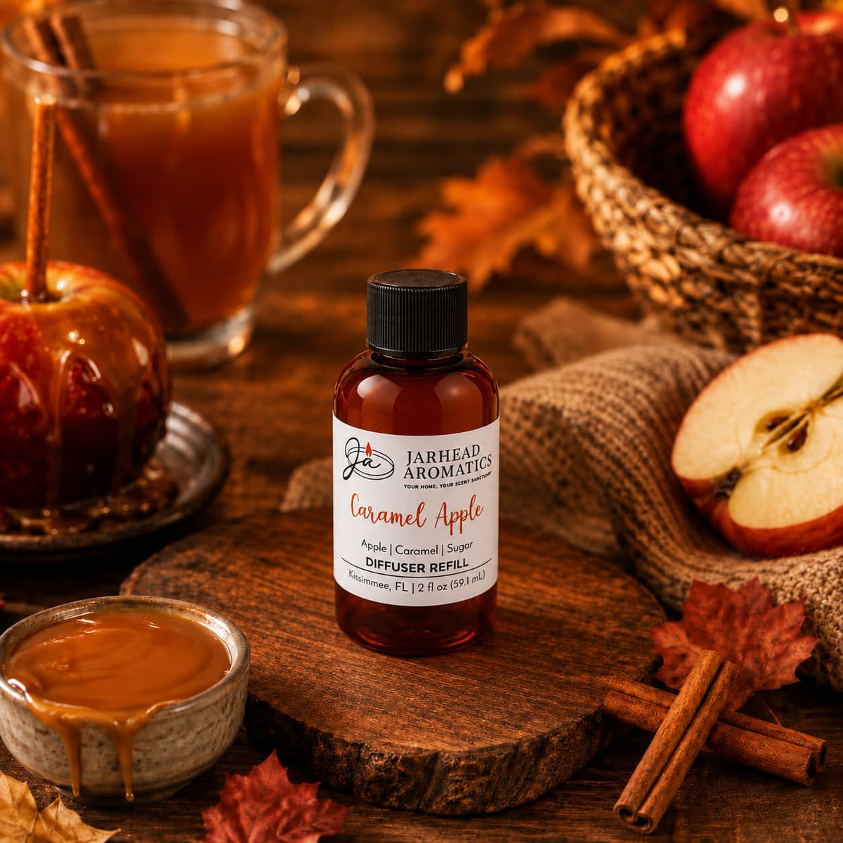 A bottle of Jarhead Aromatics Caramel Apple Diffuser Refill | Crisp Apple, Rich Caramel & Warm Spice is surrounded by caramel, apples, cinnamon sticks, and autumn leaves, with a mug and a basket of apples in the background. This cozy autumn-themed arrangement beautifully captures the inviting essence of the scent—perfect for elevating your home fragrance.