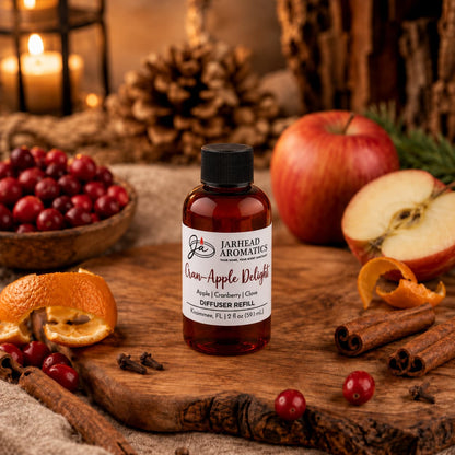 A bottle of Cran-Apple Delight Diffuser Refill | Cranberry, Apple & Warm Cinnamon by Jarhead Aromatics sits on a rustic wooden surface, surrounded by fresh apples, tart cranberries, vibrant orange peels, cinnamon sticks, and aromatic cloves. The inviting home fragrance scene is enhanced with candles and pinecones in the background.