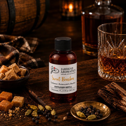 A bottle of Jarhead Aromatics Sweet Bourbon Diffuser Refill—featuring notes of bourbon, butterscotch, and brown sugar—rests on a table alongside a glass of bourbon, sugar cubes, cardamom pods, peppercorns, cinnamon sticks, and a plaid cloth, setting a warm and rustic home fragrance ambiance.