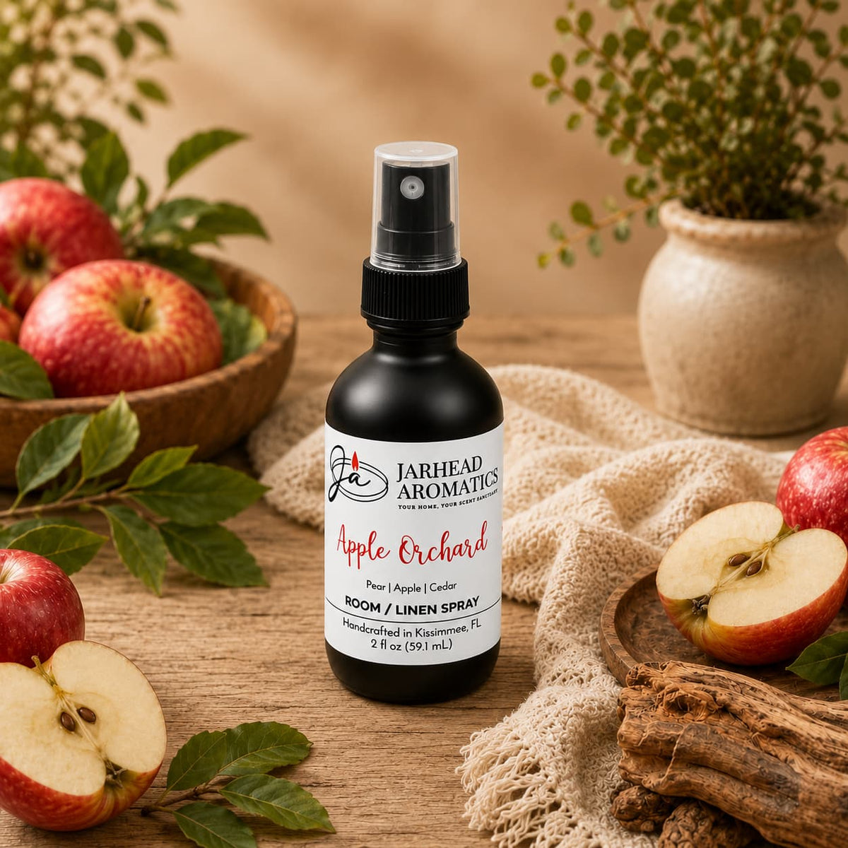 A bottle of Jarhead Aromatics Apple Orchard Room & Linen Spray | Crisp Apple, Pear & Cedarwood sits on a table surrounded by fresh apples, apple slices, greenery, and a woven basket in a rustic setting, perfectly capturing the essence of this premium home fragrance spray for both rooms and linens.